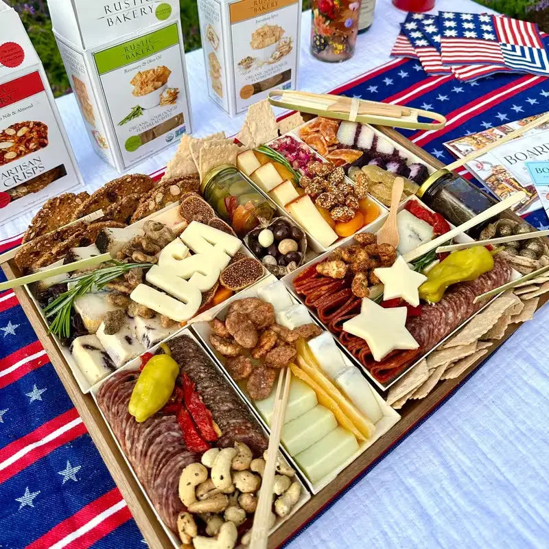 Patriotic-themed charcuterie board with assorted cheeses, meats, nuts, & crackers, featuring cheese cutouts shaped like "USA" & stars