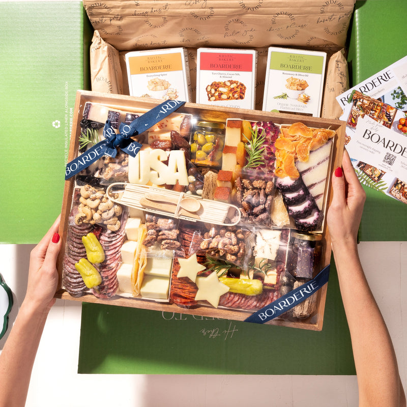 A Boarderie Labor Day Large USA charcuterie board gift box is held open by two hands, showcasing a curated assortment of cheeses, crackers, cured meats, nuts, and garnishes.