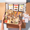 Hands holding an large charcuterie gift box with assorted cheeses, nuts, and dried fruits, packaged with artisan crisps & Boarderie brochures