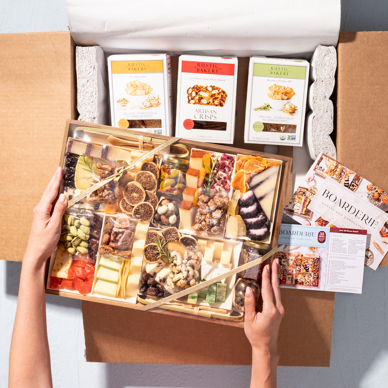 Hands holding an large charcuterie gift box with assorted cheeses, nuts, and dried fruits, packaged with artisan crisps & Boarderie brochures