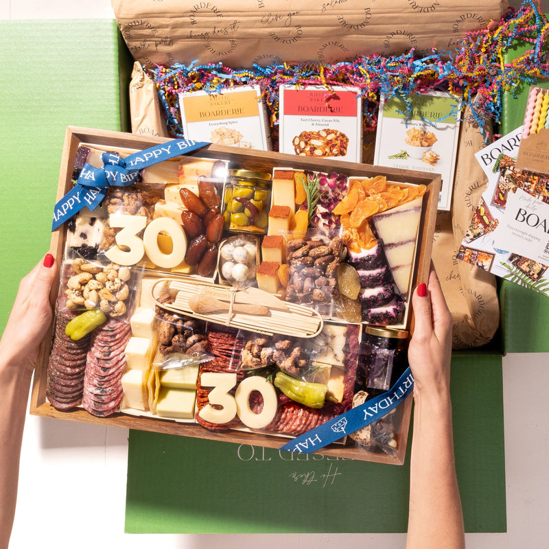 Gourmet Boarderie Cheese & Charcuterie Board with 'Happy Birthday' and '30' decorations, held by two hands.