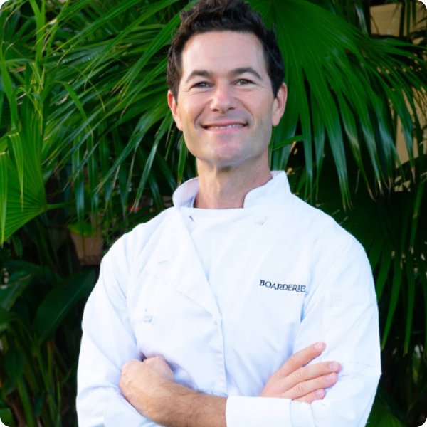 Aaron Menitoff in white uniform smiling with arms crossed in front of lush green palm leaves.