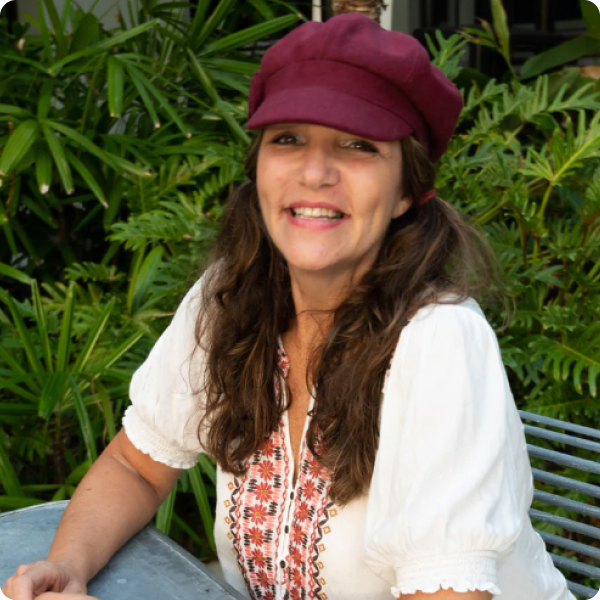 Julie Menitoff wearing a burgundy cap and embroidered blouse, seated outdoors among green plants.