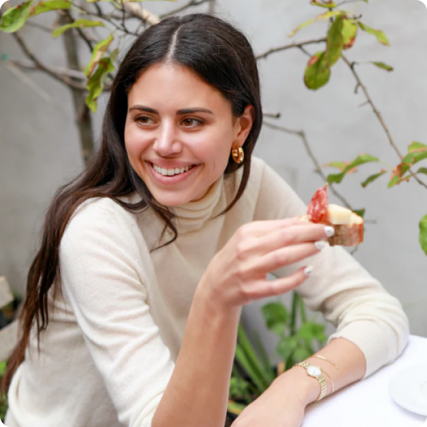 Rachel Solomon smiling while holding a piece of bread topped with cheese and charcuterie, seated outdoors.