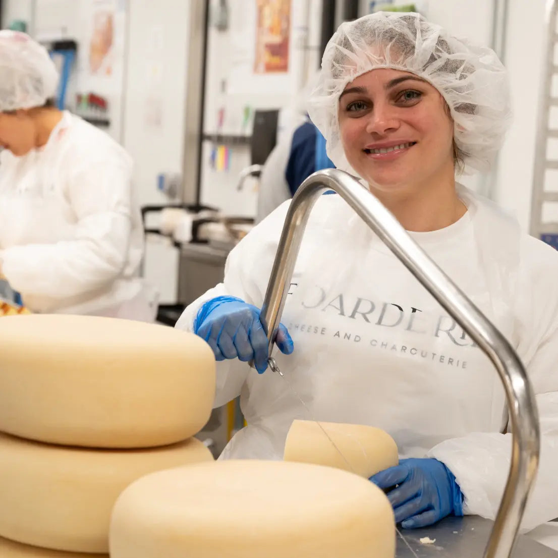Boarderie cheesemaker cutting large wheels of cheese in a processing room wearing protective gear.
