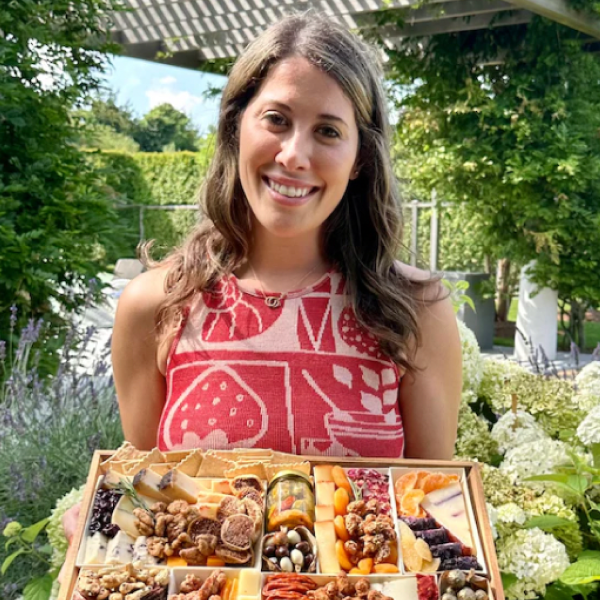 Lanie Shalek smiling and holding a charcuterie board filled with cheeses, nuts, and fruits in an outdoor garden setting.