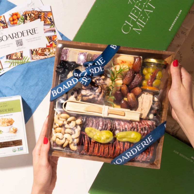 Artisan cheese & charcuterie boards with gourmet meats, cheeses, nuts, dried fruits, olives, and rosemary garnish in gift packaging.