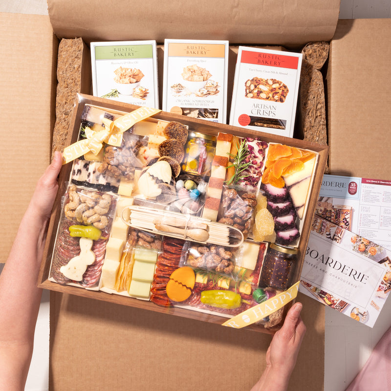 Hands holding an Easter-themed charcuterie box with cheeses, meats, and festive decorations, wrapped with a "Happy Easter" ribbon.