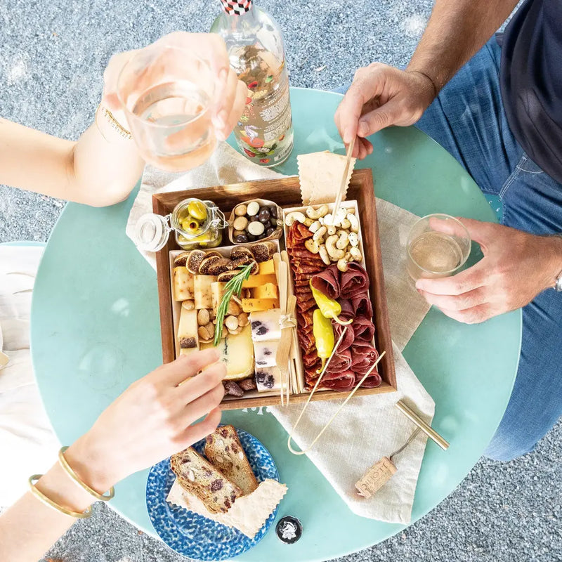 Two people enjoying a charcuterie board with cheese, meats, nuts, & drinks at an outdoor table