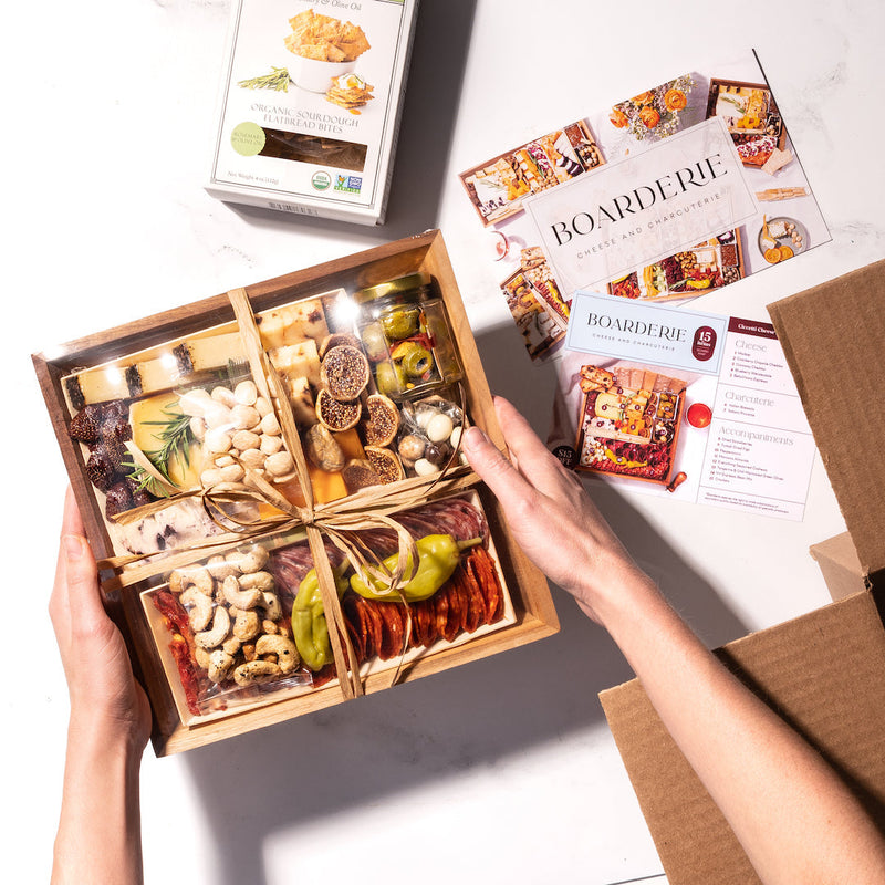 Hands holding a small charcuterie gift box with assorted cheeses, meats, nuts, & figs, alongside Boarderie pamphlets on a white background