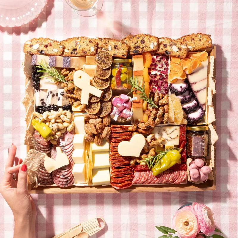 Charcuterie board with cheeses, meats, dried fruits, and heart-shaped cheese slices on a pink gingham tablecloth.