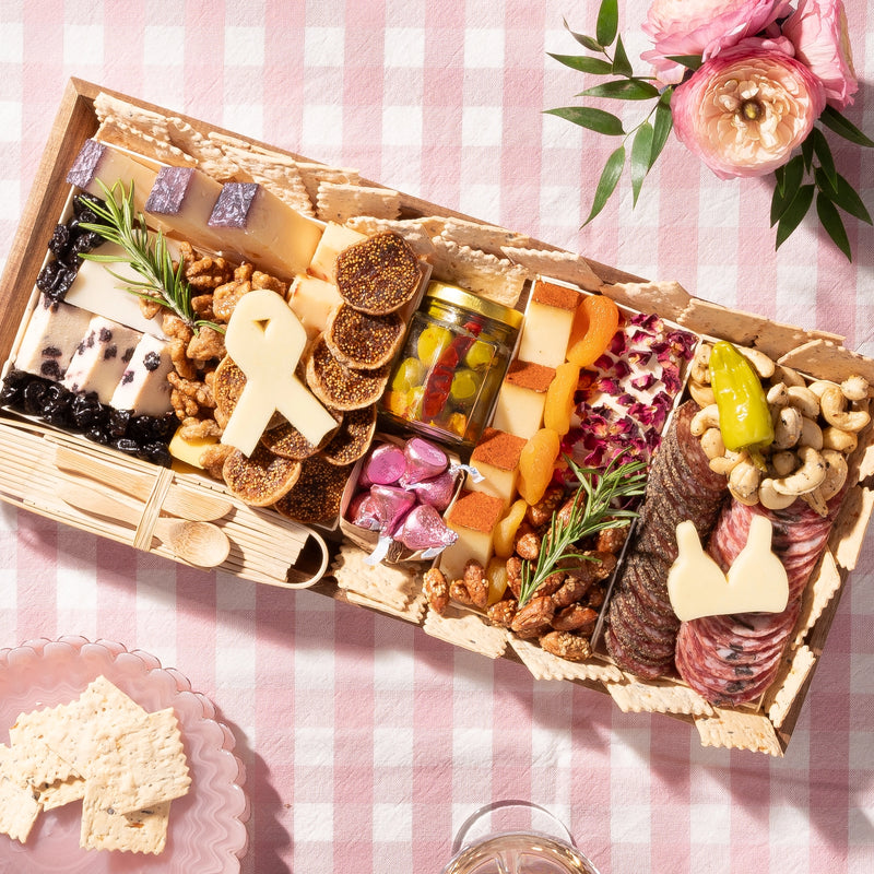 large Charcuterie board with cheeses, meats, nuts, & a ribbon-shaped cheese piece, arranged on a pink checkered tablecloth with roses on the side