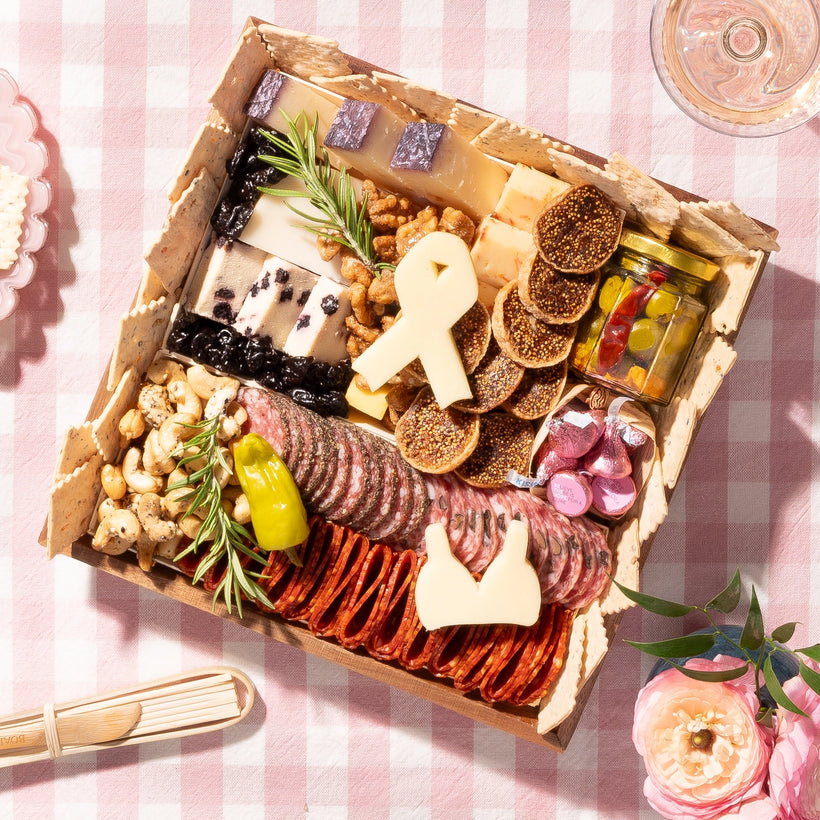 Charcuterie board with cheeses, meats, nuts, & a ribbon-shaped cheese piece, arranged on a pink checkered tablecloth with decorative accents