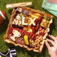 Super Bowl-themed charcuterie board with cheeses, meats, crackers, nuts, and football-inspired cheese cutouts on a grass picnic setting.