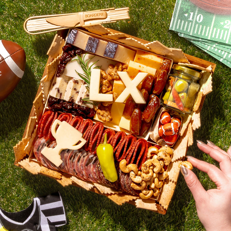 Super Bowl-themed charcuterie board with cheeses, meats, crackers, nuts, and football-inspired cheese cutouts on a grass picnic setting.