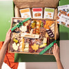 Hands holding a Thanksgiving charcuterie board featuring cheeses, nuts, dried fruits, & leaf-shaped cheese pieces, wrapped with a green Boarderie ribbon