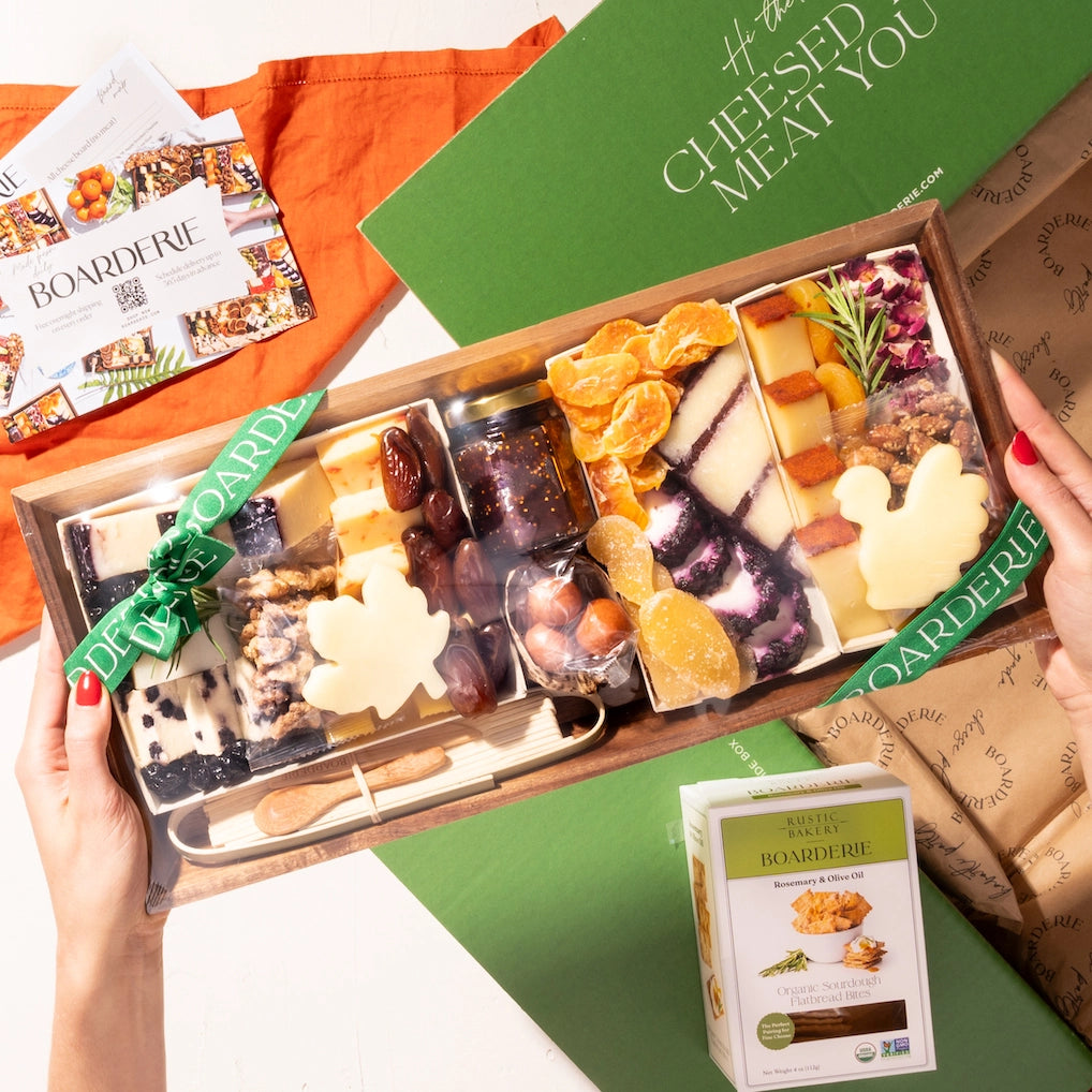 Hands holding a fall-themed charcuterie board featuring cheeses, dried fruit, nuts, & leaf-shaped cheese slices, wrapped in a green Boarderie ribbon