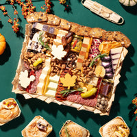 An overhead styled food presentation showing a large charcuterie board filled with cheeses, cured meats, nuts, crackers, fruit, and garnishes, surrounded by small baked goods and seasonal pumpkins on a dark green background.