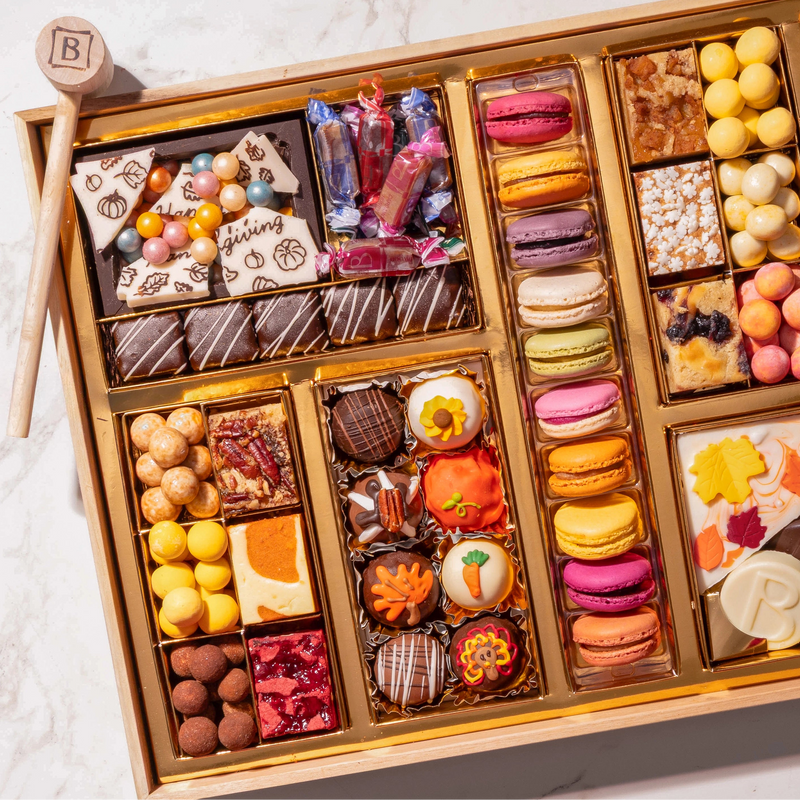 Close-up of Boarderie’s Thanksgiving Large Dessert Board showing the smashed chocolate box with chocolate pearls and Thanksgiving bark shards, rows of seasonal macarons in Pumpkin Spice, Apple Pie, and Sweet Potato Pie flavors, hand-painted autumn truffles, Pumpkin Cheesecake Blondie, Lemon Blueberry Blondie, Red Velvet Brownie, toffee, and Creme Brulee Cordials on an acacia wood board