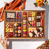 Overhead view of Boarderie’s Thanksgiving Large Dessert Board with 47+ artisan confections including a Happy Thanksgiving chocolate smash box, wrapped butter caramels, colorful seasonal macarons, hand-painted turkey, pumpkin, and leaf truffles, Thanksgiving White Chocolate Cheesecake Bark, chocolate-dipped Oreos, brownies, blondies, toffee, chocolate-covered pretzels, and sweet bites in gold boxes on a reusable acacia wood board