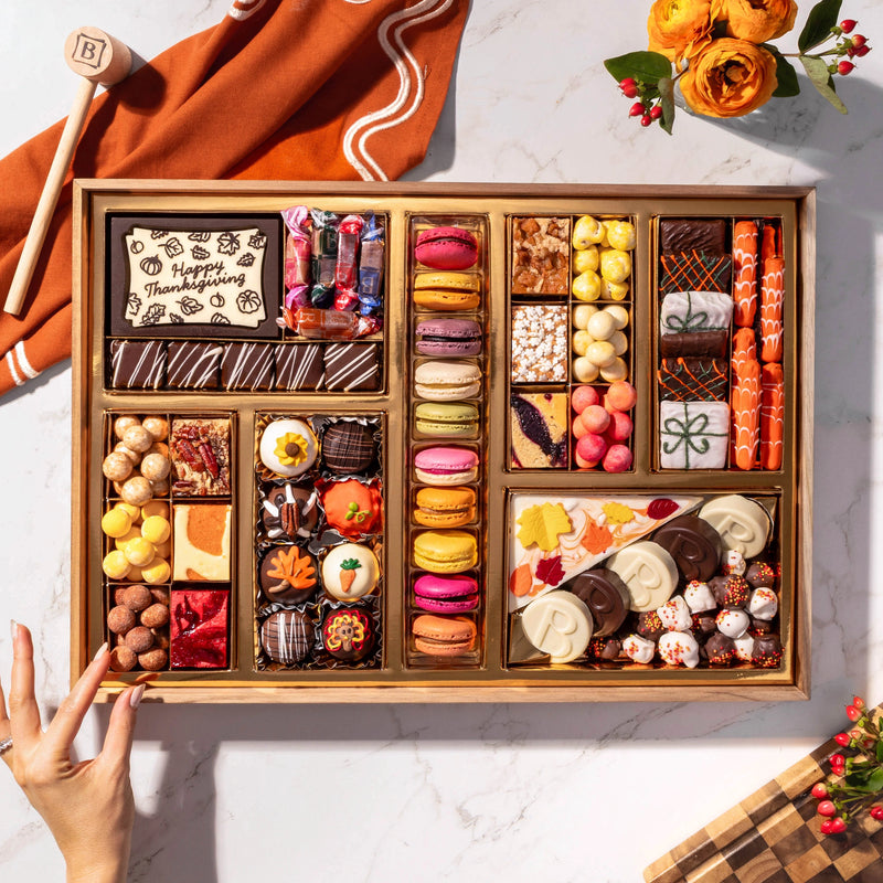 Overhead view of Boarderie’s Thanksgiving Large Dessert Board with 47+ artisan confections including a Happy Thanksgiving chocolate smash box, wrapped butter caramels, colorful seasonal macarons, hand-painted turkey, pumpkin, and leaf truffles, Thanksgiving White Chocolate Cheesecake Bark, chocolate-dipped Oreos, brownies, blondies, toffee, chocolate-covered pretzels, and sweet bites in gold boxes on a reusable acacia wood board