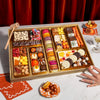 Boarderie Thanksgiving Large Dessert Board on a marble table with a guest reaching for a chocolate truffle, showing the full 47+ piece dessert spread with macarons, Thanksgiving White Chocolate Cheesecake Bark, Boarderie-branded dark and white chocolate-dipped Oreos, caramel popcorn bites, wrapped butter caramels, and the wooden smash hammer, styled with champagne glasses, fall decor, and orange linen napkins