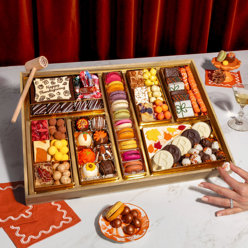 Boarderie Thanksgiving Large Dessert Board on a marble table with a guest reaching for a chocolate truffle, showing the full 47+ piece dessert spread with macarons, Thanksgiving White Chocolate Cheesecake Bark, Boarderie-branded dark and white chocolate-dipped Oreos, caramel popcorn bites, wrapped butter caramels, and the wooden smash hammer, styled with champagne glasses, fall decor, and orange linen napkins