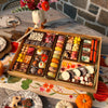 Boarderie Thanksgiving Large Dessert Board displayed on a rustic outdoor dining table set for a Thanksgiving gathering with fall flowers, pumpkins, wine glasses, and a floral tablecloth, showing the full spread of 47+ artisan desserts including macarons, truffles, cheesecake bark, chocolate-dipped Oreos, and caramel popcorn bites
