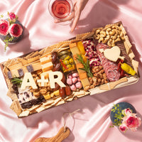 Luxury Valentine’s Day charcuterie board gift by Boarderie featuring artisan cheeses, cured meats, dried fruits, nuts, crackers, gourmet spreads, and customized initials in cheese. Board is on a pink romantic backdrop, styled for nationwide overnight gifting. 