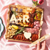 Luxury Valentine’s Day charcuterie board gift by Boarderie featuring artisan cheeses, cured meats, dried fruits, nuts, crackers, gourmet spreads, and customized initials in cheese. Board is on a pink romantic backdrop, styled for nationwide overnight gifting. 