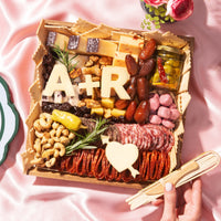 Luxury Valentine’s Day charcuterie board gift by Boarderie featuring artisan cheeses, cured meats, dried fruits, nuts, crackers, gourmet spreads, and customized initials in cheese. Board is on a pink romantic backdrop, styled for nationwide overnight gifting. 