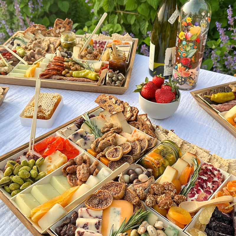 Assorted charcuterie boards with cheeses, nuts, and fruits set on a white tablecloth with wine bottles in an outdoor garden setting.