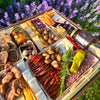 close-up of a charcuterie board featuring meats, cheeses, nuts, dried fruits, & a jar of preserves, set outdoors