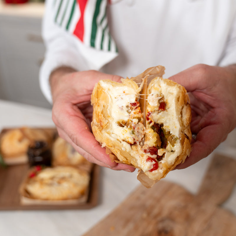 Hands holding a savory baked brie pastry split open, showing melted cheese, sun-dried tomatoes, and herbs.