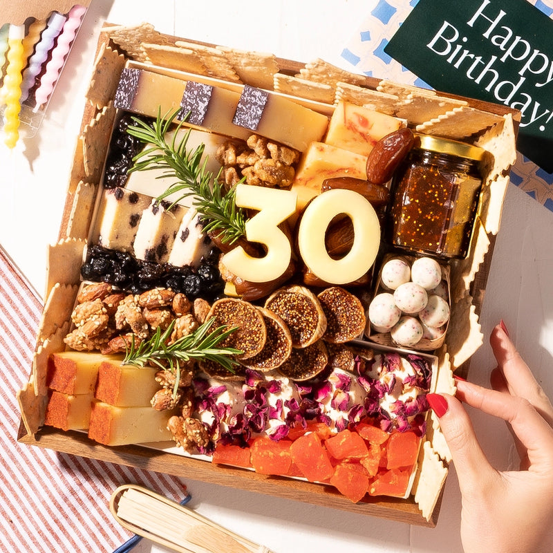 small birthday-themed charcuterie board gift featuring cheeses, nuts, figs, fruit, crackers, & a "30" made of cheese