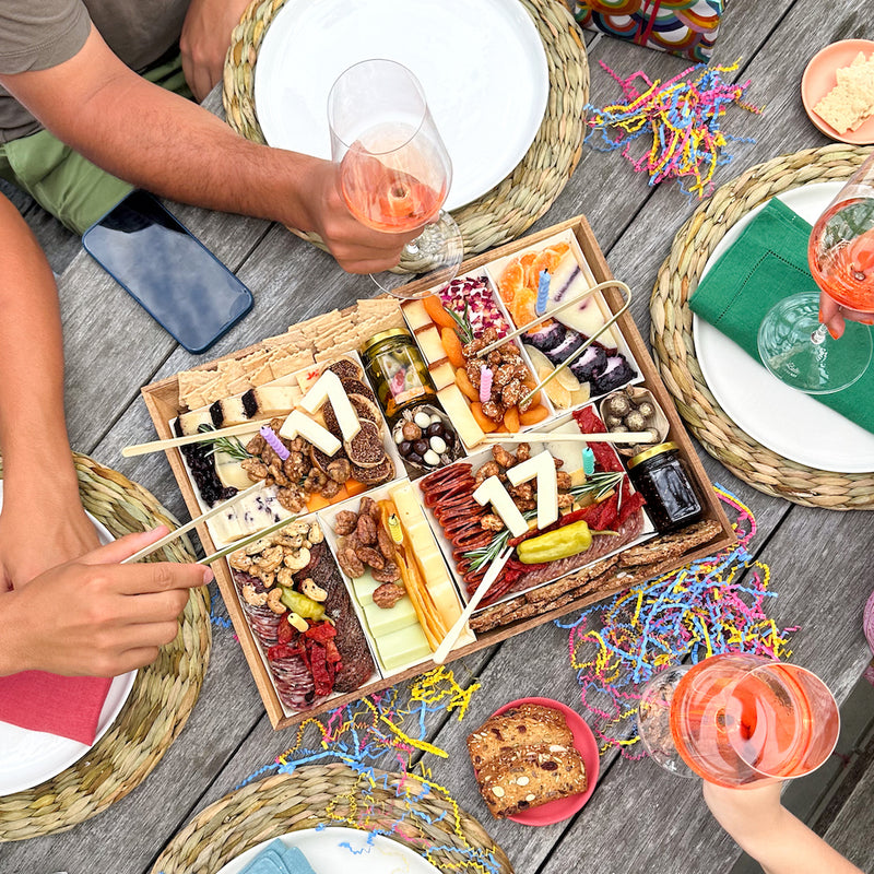 Charcuterie board with "17" in cheese, surrounded by people holding wine glasses at an outdoor celebration table.