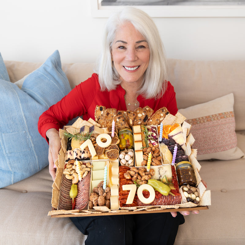 Smiling woman holding a birthday charcuterie board with "70" cheese numbers, featuring a variety of meats, cheeses, and colorful candles.