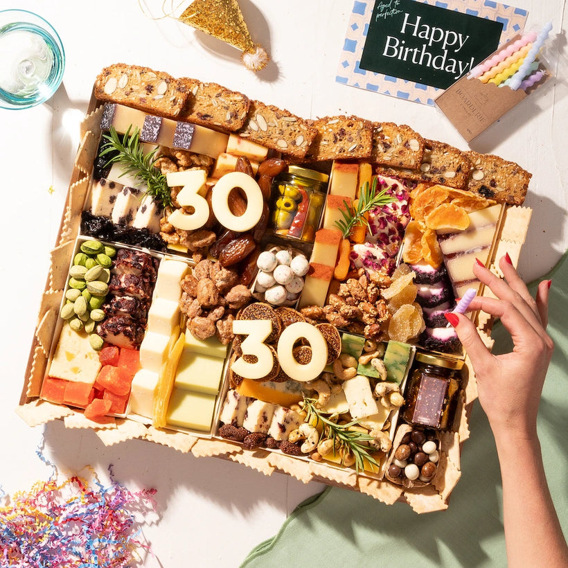 Birthday cheese board featuring an assortment of cheeses, nuts, dried fruits, and "30" cheese numbers, with festive decorations and candles.