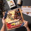 Hands holding a gourmet charcuterie box with cheeses, almonds, truffle hot sauce, & other delicacies, wrapped with a Boarderie ribbon