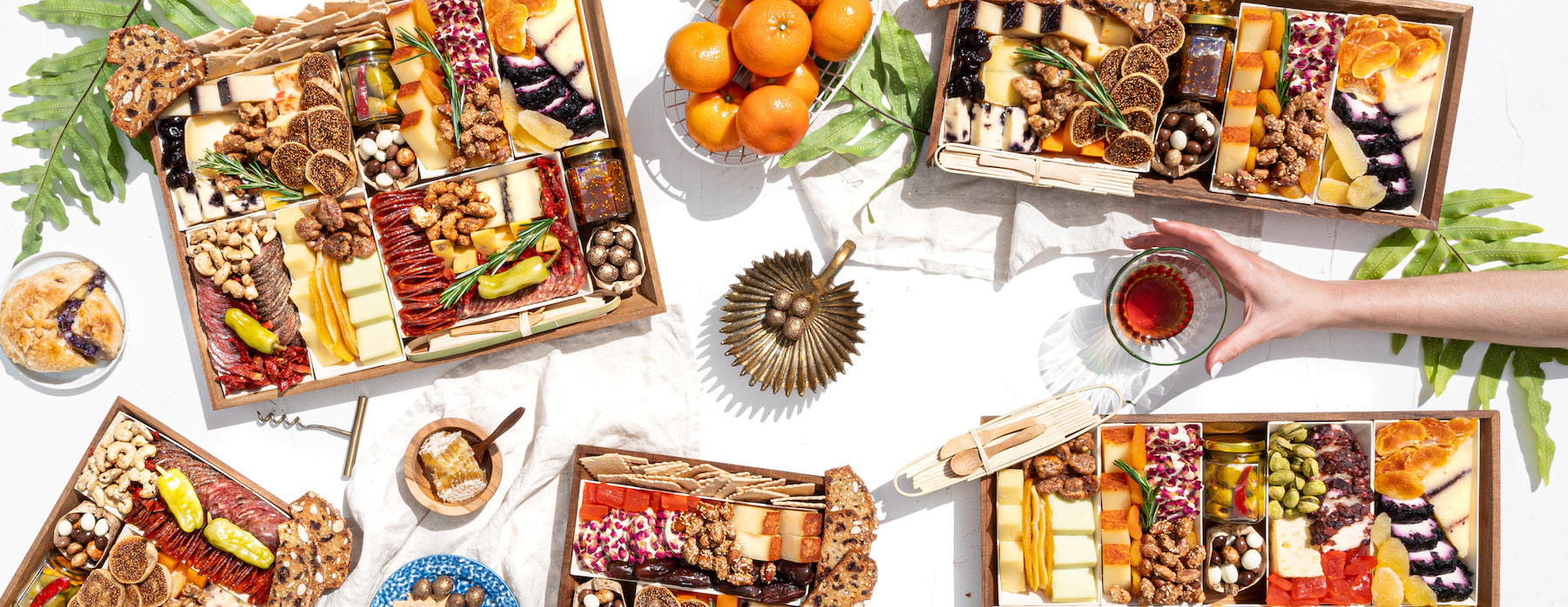 Assorted charcuterie boards with cheeses, meats, nuts, and fruits arranged on a table with a hand reaching for a drink.