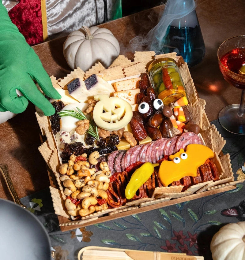 Halloween-themed charcuterie board with jack-o'-lantern cheese cutout, googly-eyed candy treats, cured meats, nuts, olives, and white decorative pumpkin.