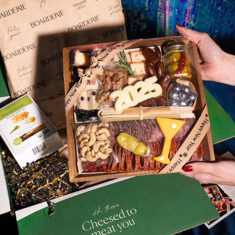 charcuterie board with new year's 2025 theme being held up out of box. Board features an assortment of cheeses, nuts, dried fruits, & olives, wrapped in a happy new year ribbon