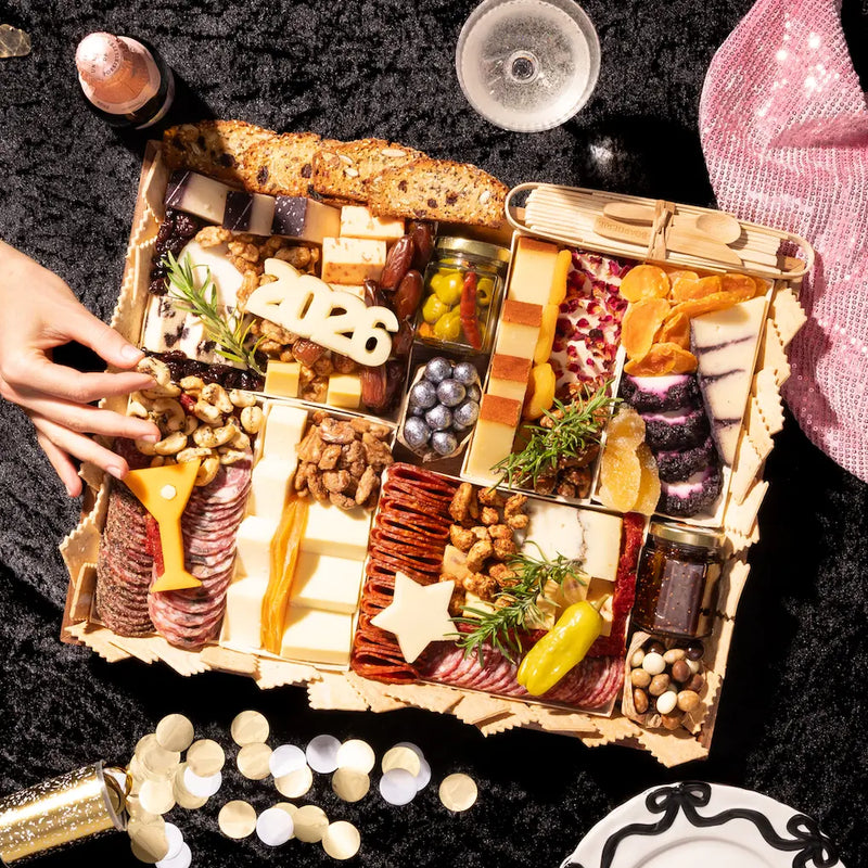 Overhead view of large New Years charcuterie board with assorted cheeses, cured meats, nuts, olives, bread, and star-shaped cheese cutouts surrounded by champagne and decorations.