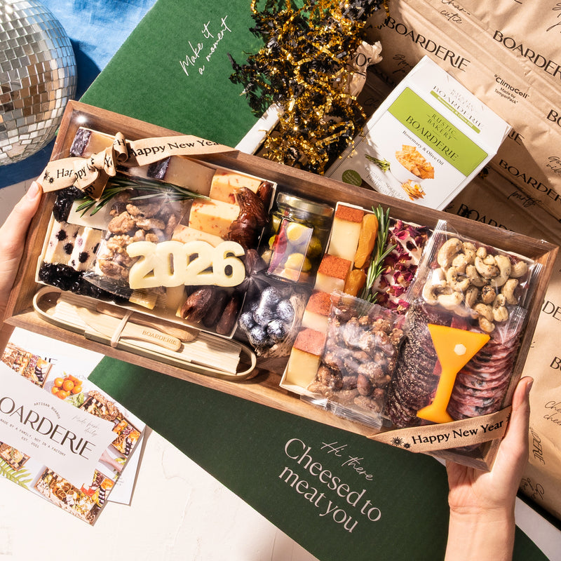 Medium New Year's  charcuterie board with "2026" cheese cutout, assorted cheeses, cured meats, nuts, jams, and serving utensils surrounded by party decorations.