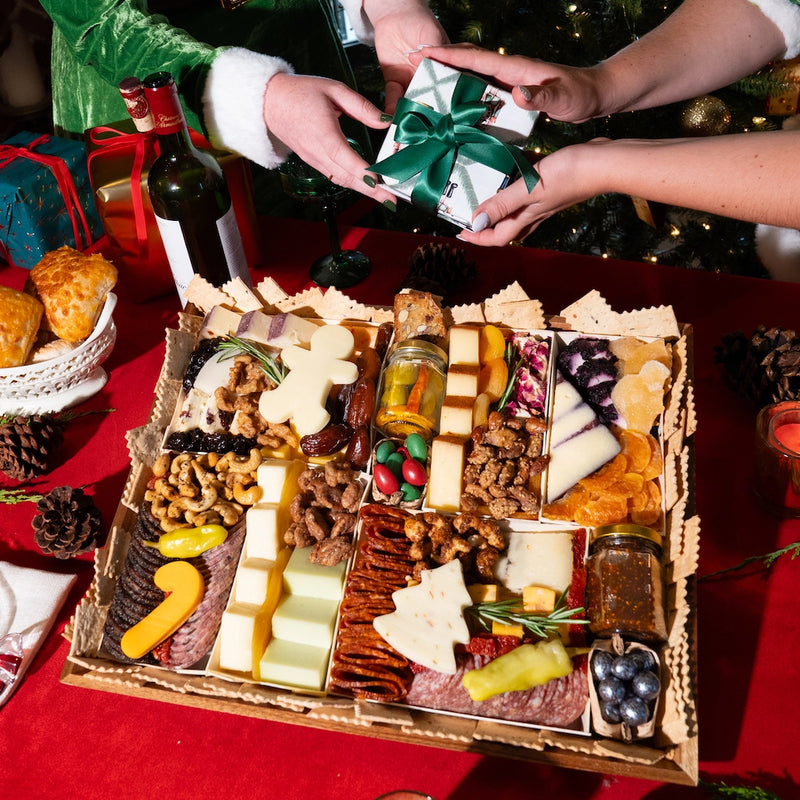 Christmas charcuterie board displayed with festive cheeses, meats, nuts, fruits, and holiday treats as gifts are exchanged nearby.