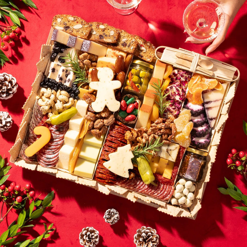 large Festive charcuterie board with holiday-themed cheeses, meats, and nuts on a red background with pinecones & holly