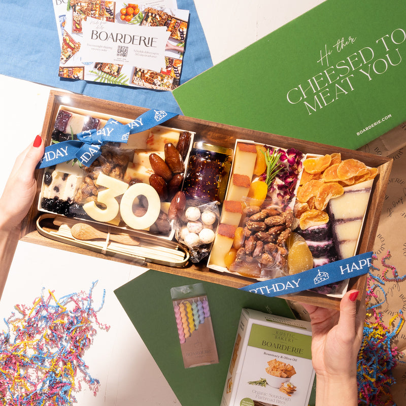 Birthday charcuterie box featuring assorted cheeses, nuts, dried fruits, and a "30" cheese decoration, with festive packaging and confetti.