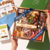 Hands holding a Boarderie charcuterie box featuring an assortment of cheeses, nuts, dried fruits, & fig spread, wrapped in branded ribbon