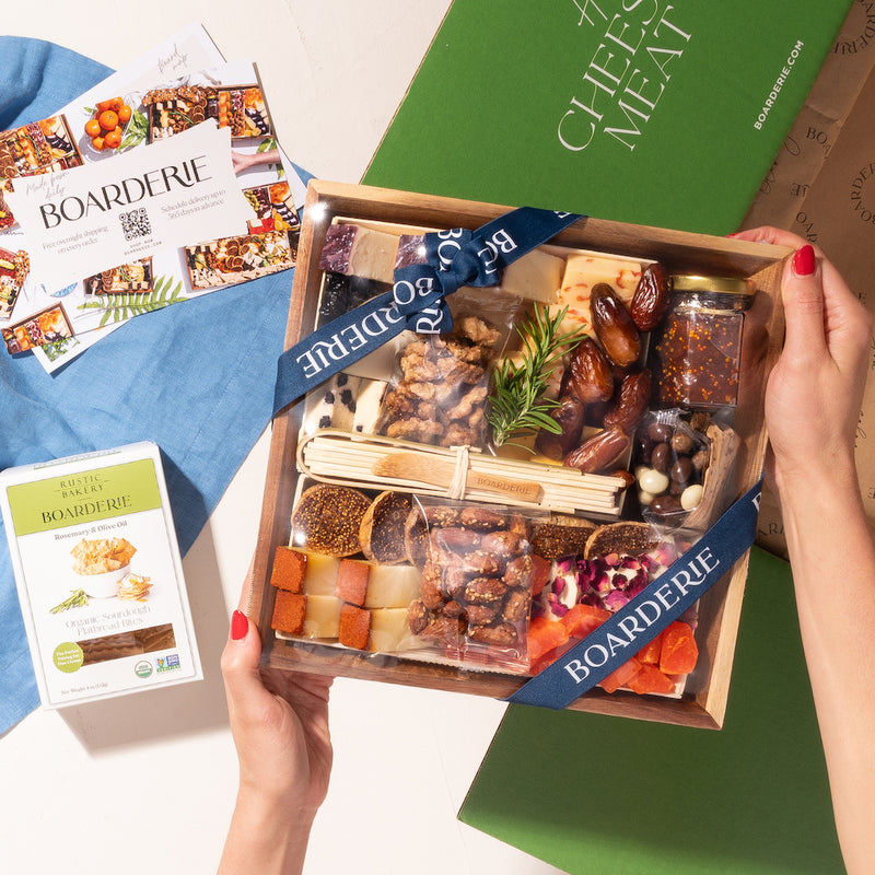 Hands holding a Boarderie charcuterie box featuring an assortment of cheeses, nuts, dried fruits, & fig spread, wrapped in branded ribbon