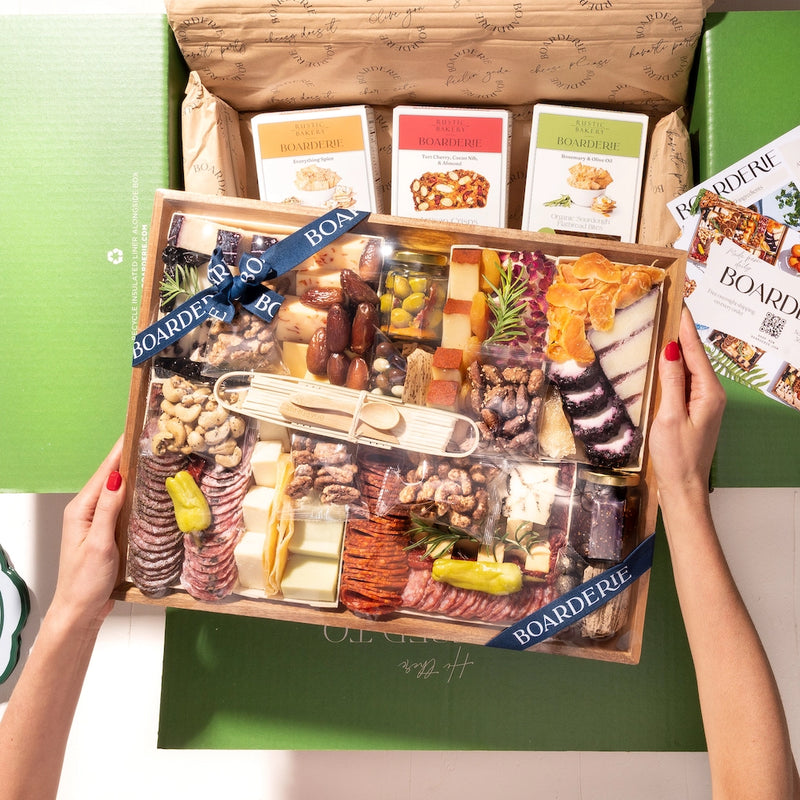 Hands holding an elaborate charcuterie board featuring cheeses, nuts, truffle hot sauce, & assorted snacks, inside a green box